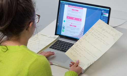 young person with long hair and glasses looking at a laptop 