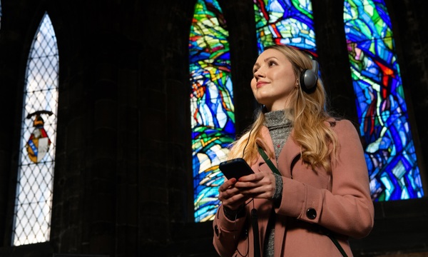 A person standing in front of a stained glass window smiling, holding an audio guide