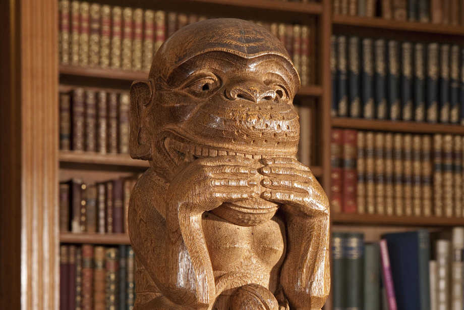 A tall wooden pole, with a carved monkey at the top, and a bookshelf standing behind the sculpture