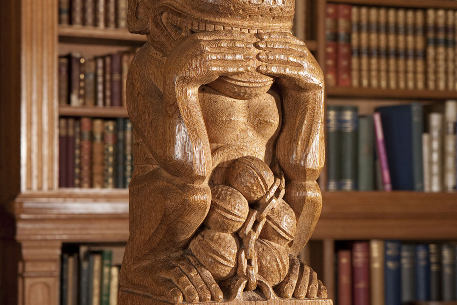 A tall wooden pole, with a carved monkey at the top, and a bookshelf standing behind the sculpture