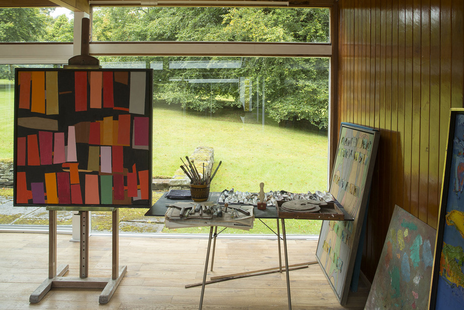 An artists workspace, with an easle, and table covered with paints and paintburshes