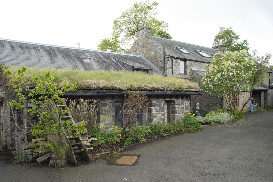 A stone farm stye building with a turf roof, beside a main building with a slate roof