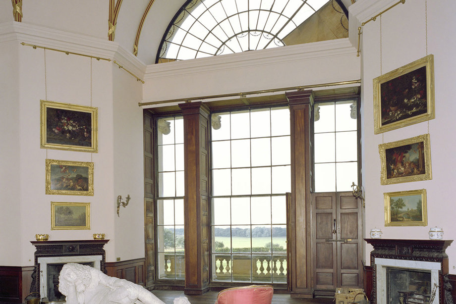 A room with large, wooden framed windows, gold-framed paintings, seating and a large stone sculpture 