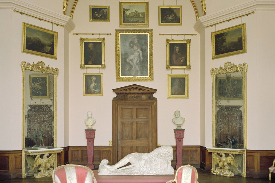 A room with a pink table with a giant sculpture on top of it, flanked by two pink and white chairs, and lots of gold-framed paintings on the walls