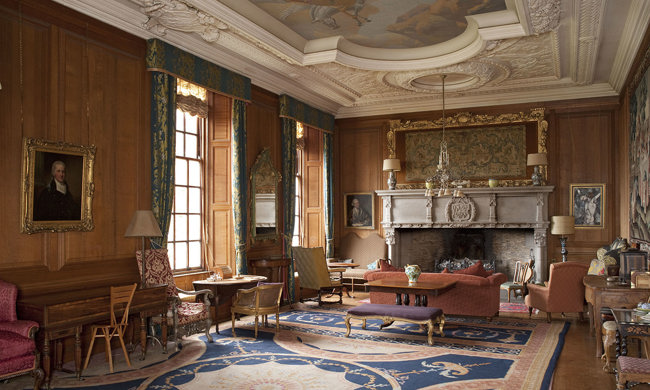 A large room with ornate plaster ceiling, wooden paneled walls and filled with furniture. A giant patterned rug covers the floor.