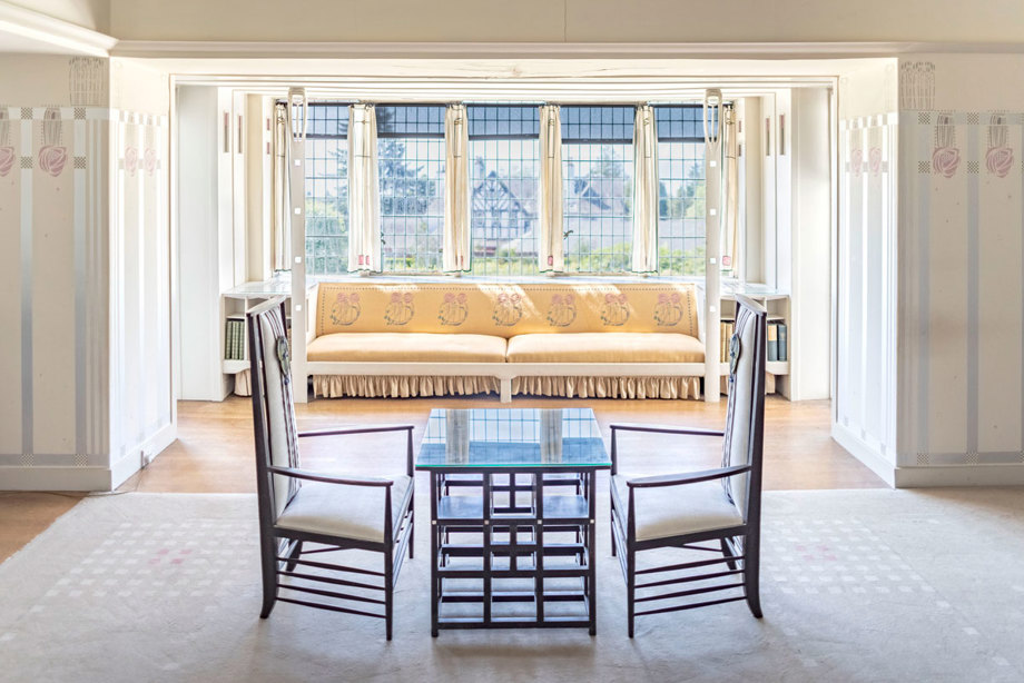 A table with two chairs in front of a large window. The window has a window seat upholstered in rose-embrioded fabric.