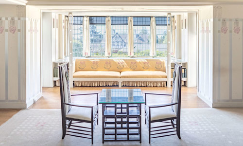 A table with two chairs in front of a large window. The window has a window seat upholstered in rose-embrioded fabric.