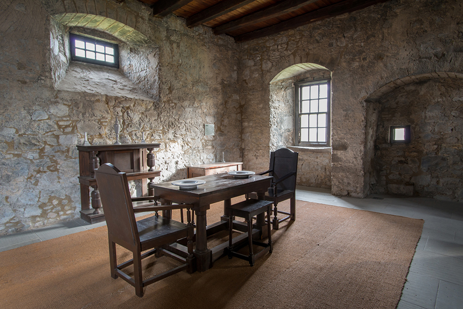 A large stone room with two windows and a fireplace. A wooden table and chairs are in the centre of the room.