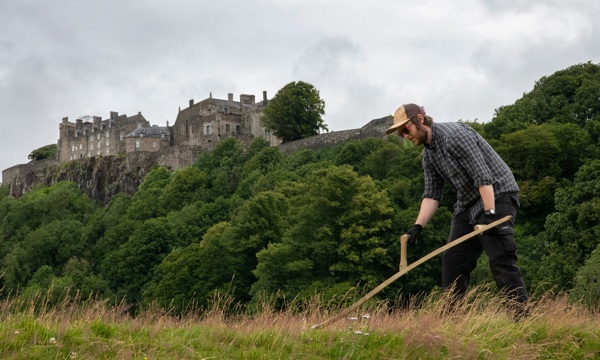 A man cutting grass with a scythe in front of Stirling Castle