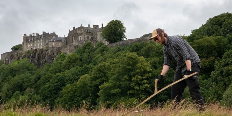 A man cutting grass with a scythe in front of Stirling Castle
