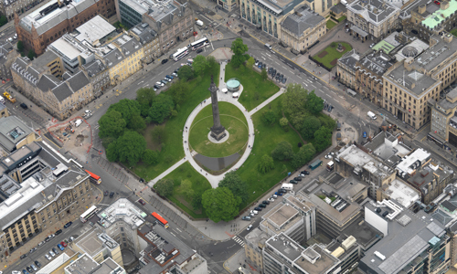 Bird's eye view of a tall monument in a green square in the centre of a city.