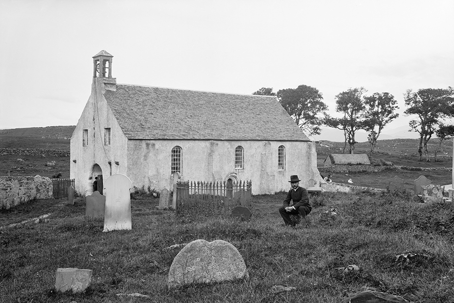 A man wearing an old fashioned suit and hat bends down in a graveyard in front of a building