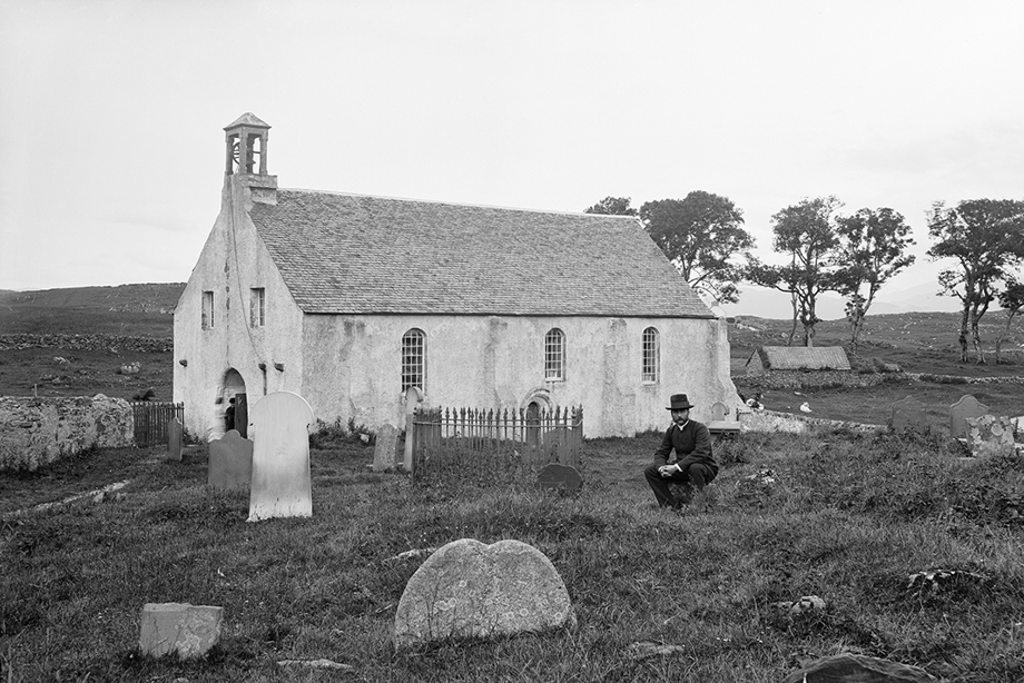 A man wearing an old fashioned suit and hat bends down in a graveyard in front of a building