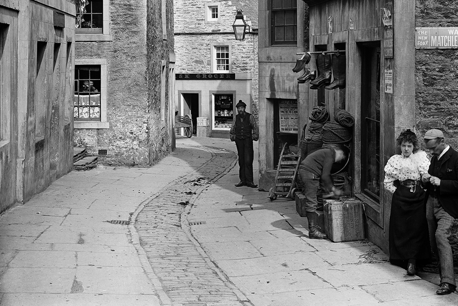A few people in historic clothing stand outside a shop where shoes and a sign reading "Private Lodgings" hang above the door