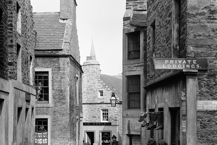 A few people in historic clothing stand outside a shop where shoes and a sign reading "Private Lodgings" hang above the door