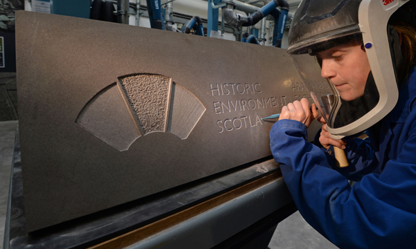 A woman wearing personal protective equipment holds a chisels a keystone into a large stone slab.