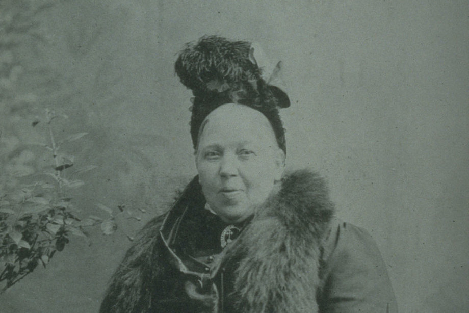 A woman in Victorian clothing and a feathered hat looks at the camera, slightly laughing