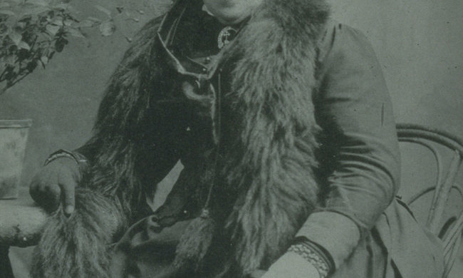 A woman in Victorian clothing and a feathered hat looks at the camera, slightly laughing