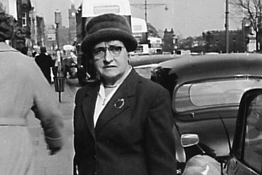 A woman holding a large handbag, wearing a hat and glasses, looking at the camera. 