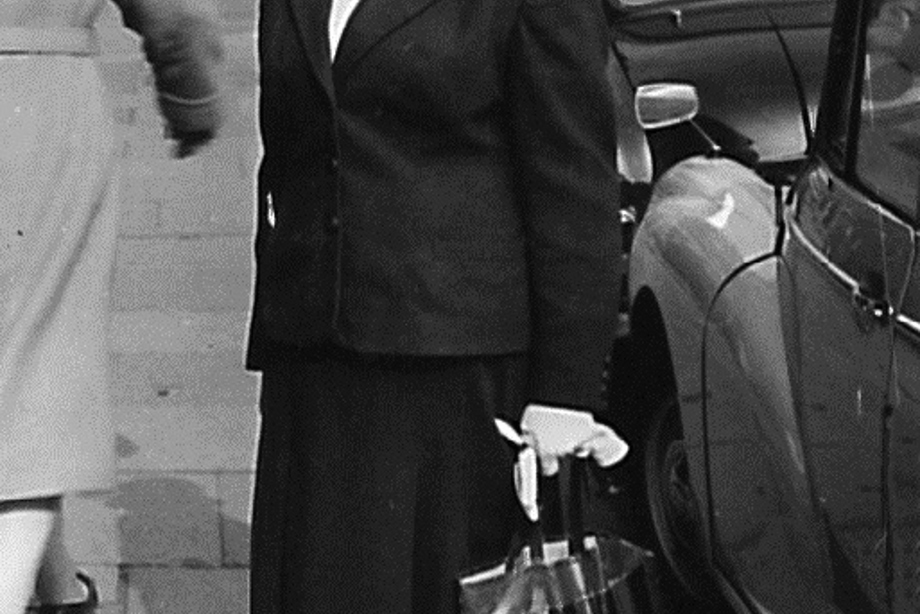 A woman holding a large handbag, wearing a hat and glasses, looking at the camera. 