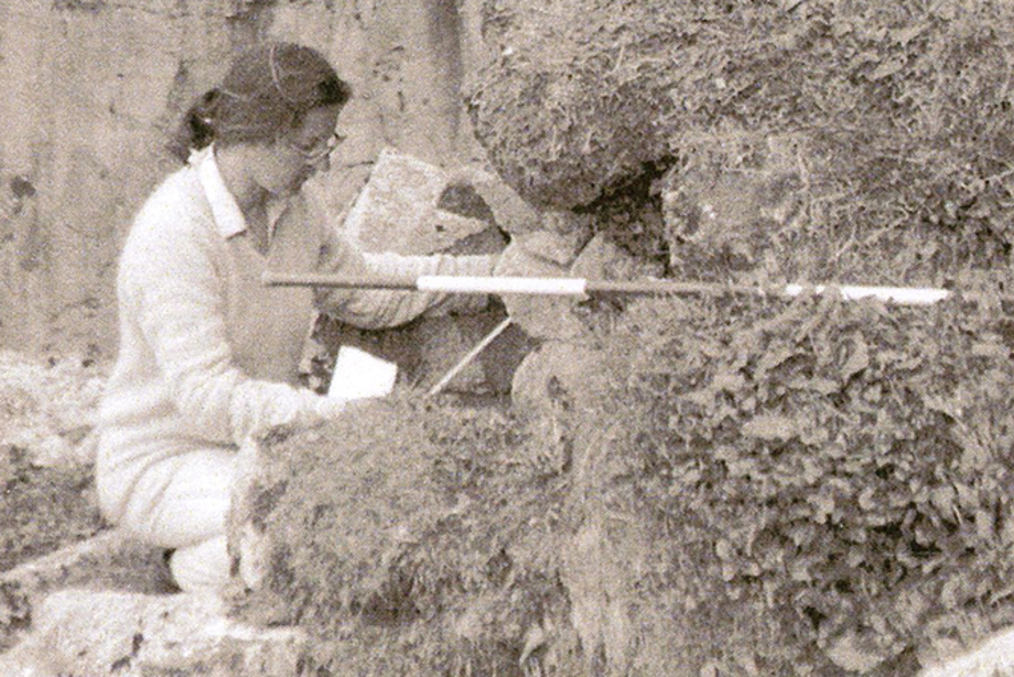 A woman wearing light coloured clothing and glasses balances a surveying tool on some rocks as she crouches 