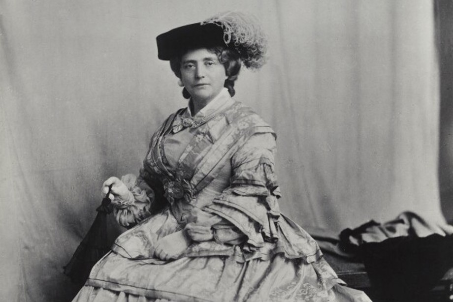 Kate Cranston wearing a large Victorian dress and a hat looks at the camera