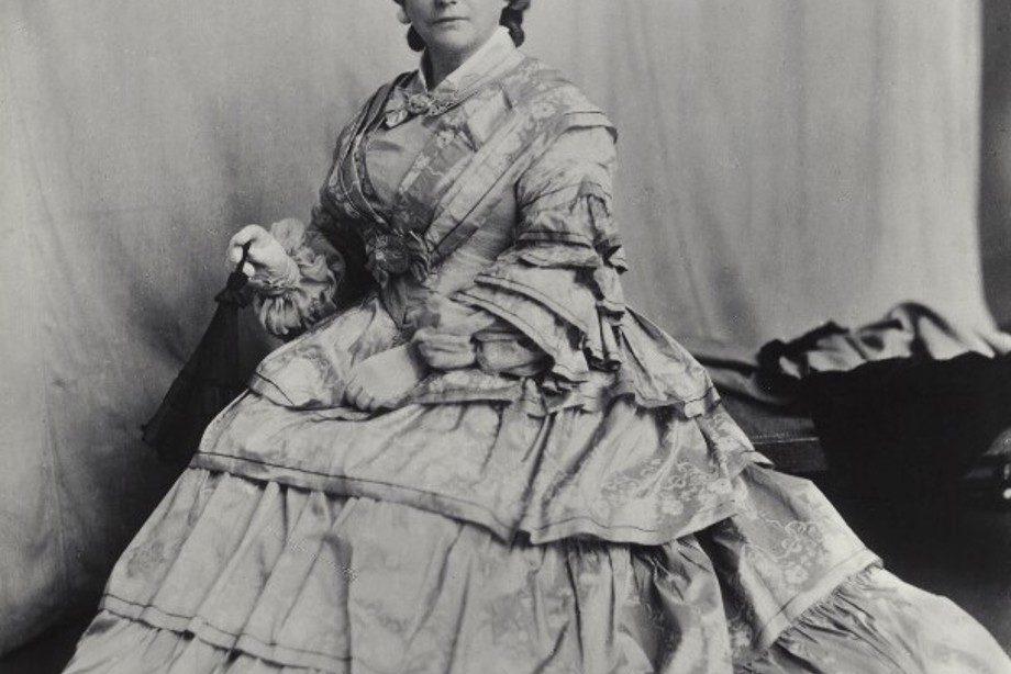 Kate Cranston wearing a large Victorian dress and a hat looks at the camera