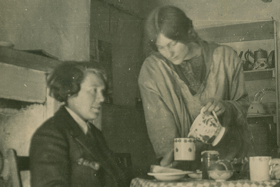 A woman pours a cup of tea for another woman who sits in a chair at the table wearing an oversized jacket