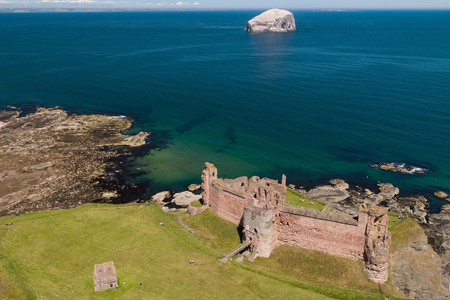 An aerial photograph of a ruined red stone castle close to the cliff face and the sea.