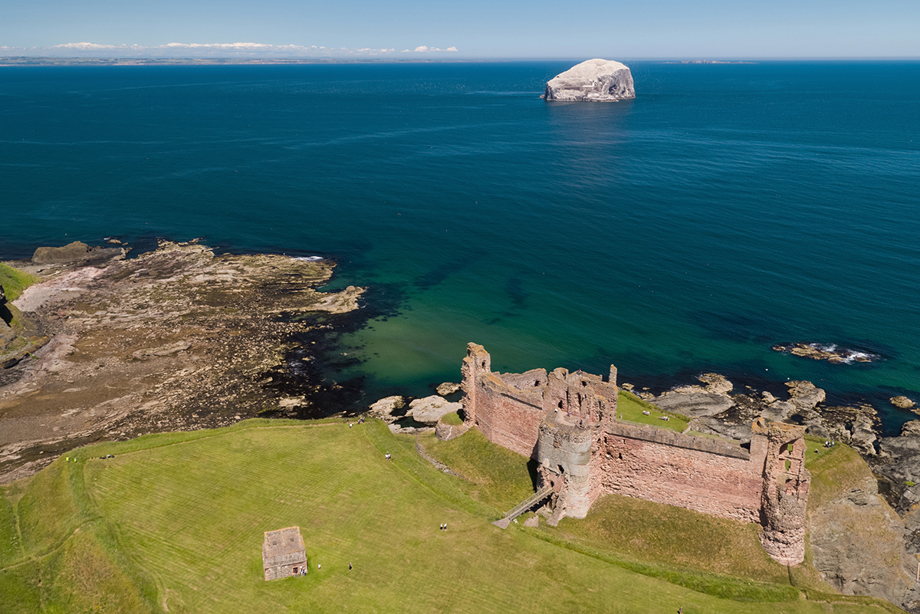 An aerial photograph of a ruined red stone castle close to the cliff face and the sea.