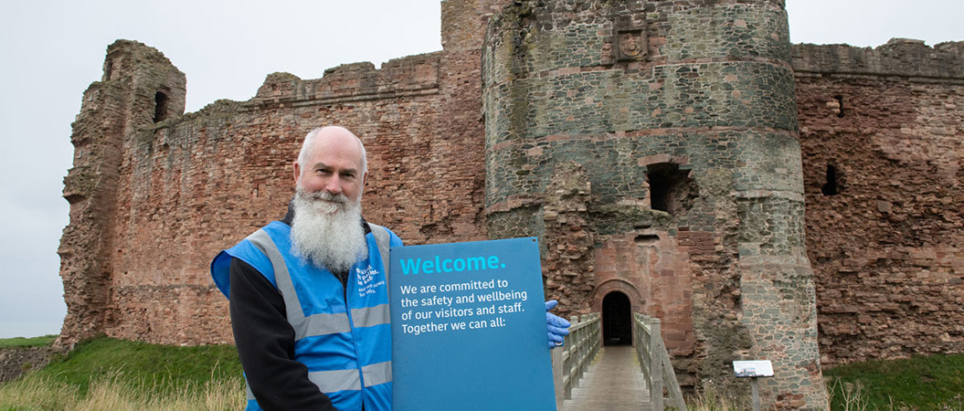 A man stands in front of Tantallon Castle, smiling, as he holds a blue board which reads "Welcome"