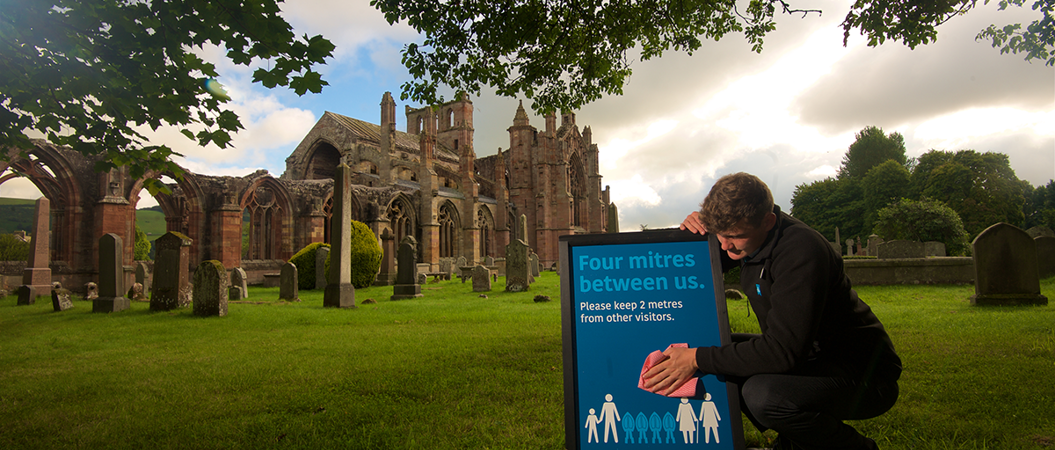 In front of a ruined abbey, a man cleans a sign which reads: "Four mitres between us"