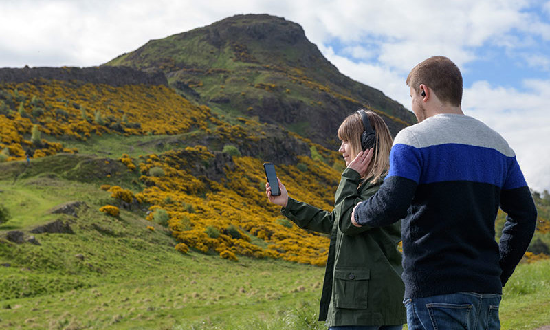 Two people standing in Holyrood Park wearing headphones and looking at a a mobile phone.
