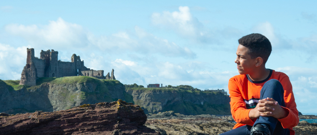 A boy sits holding his leg and looking back at Tantallon Castle