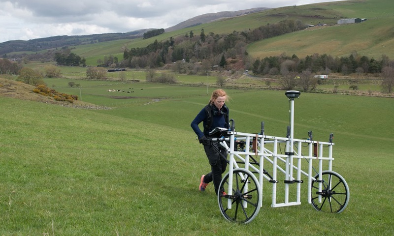 A woman pushes a large machine through a field. It looks a bit like a large metal gate on wheels.