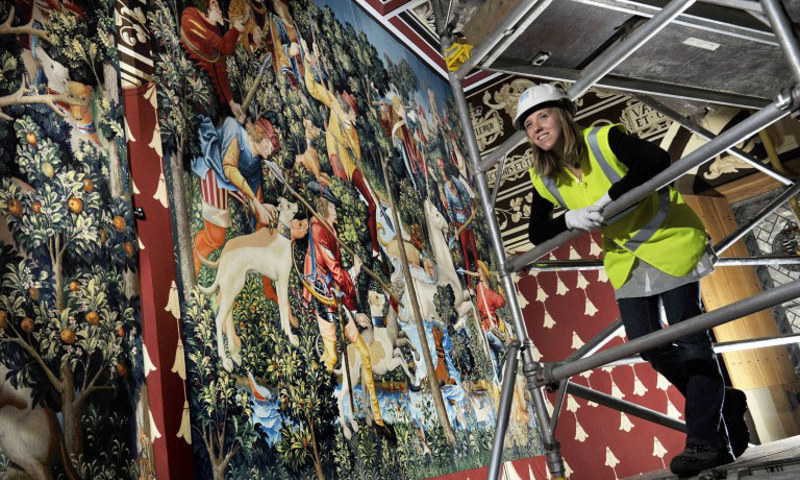a woman stands on a scaffold wearing a hard hat and high visibility vest, while se examines a large tapestry