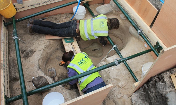 photo looking down on two archaeologists working at an excavation, lying on top of a scaffold to protect the site under them