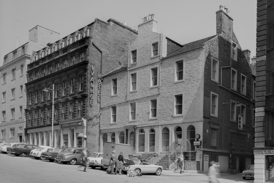 A hotel building with many windows and the words "Ivanhoe Hotel" down the side of the wall. Multiple vintage cars are parked outside.