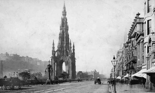 Horse drawn carriages pass by a large, gothic monument on a cobbled street