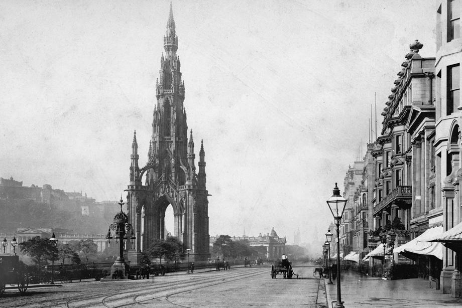 Horse drawn carriages pass by a large, gothic monument on a cobbled street