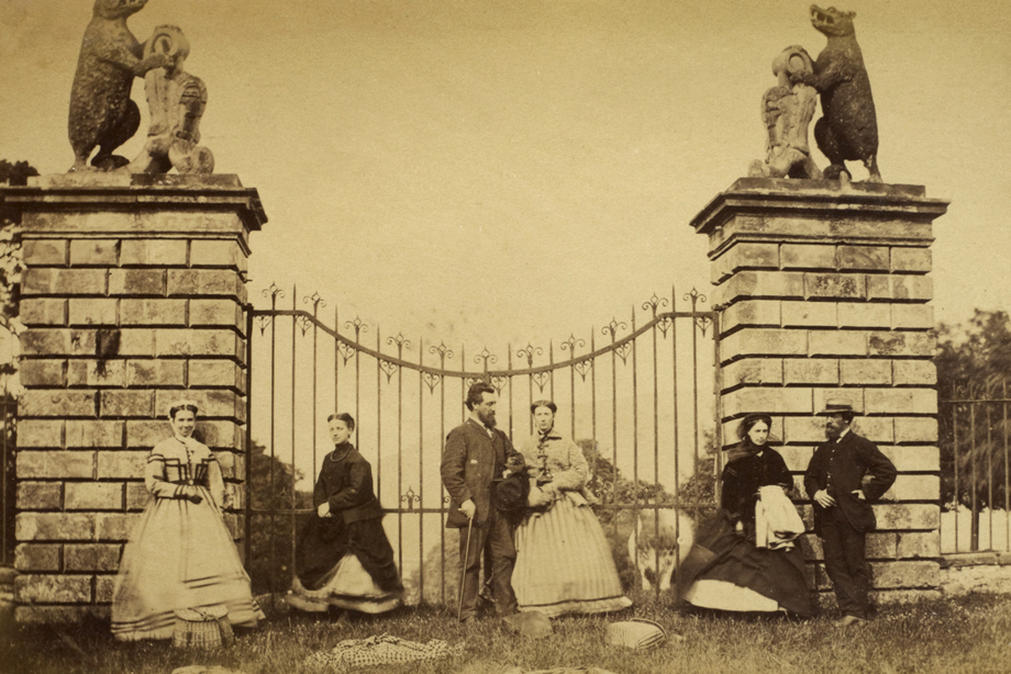 Iron gates flanked by two stone pillars featuring bear carvings and a group of people in Victorian clothing standing in front of it