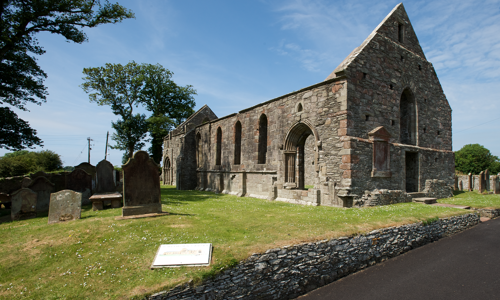 Blue skies over the roofless ruin of a stone priory building, surrounded by tombstones and green trees