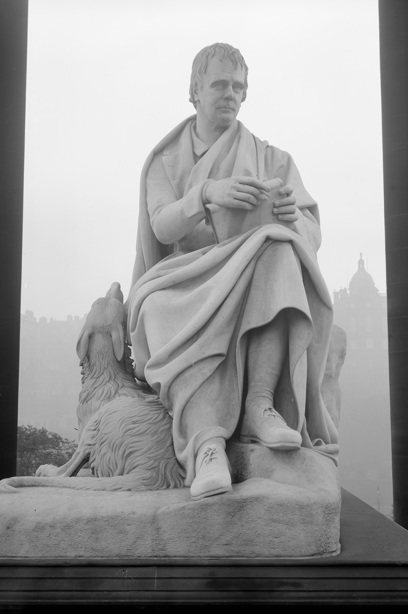 A man sits, holding a book, as his dog looks up at him