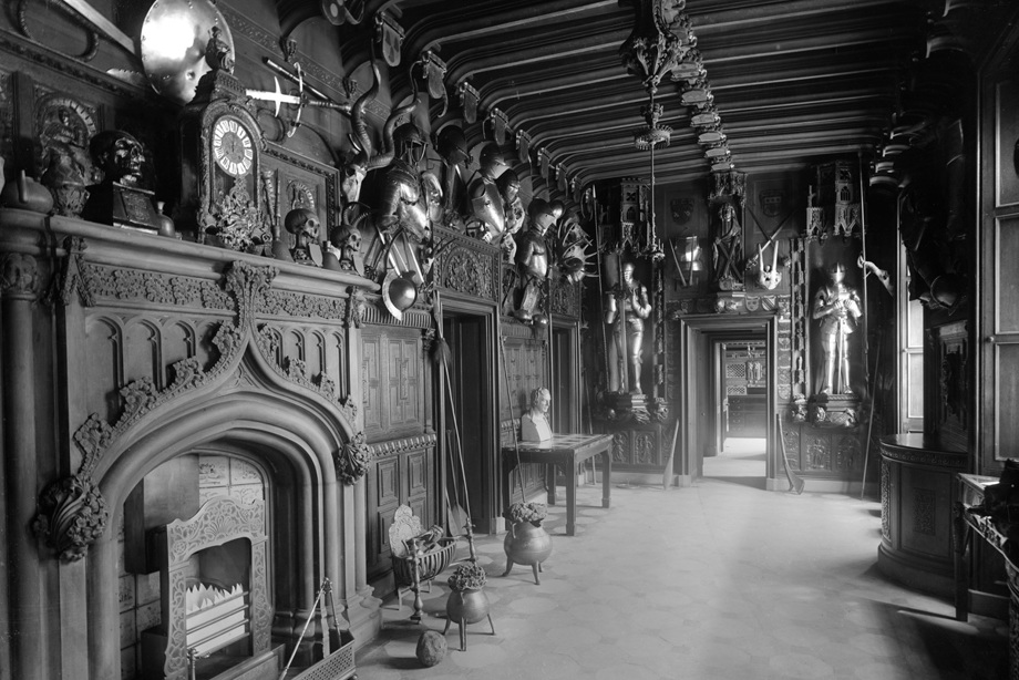 A hallway covered in wooden panelling, an intricately carved fireplace and various weapons
