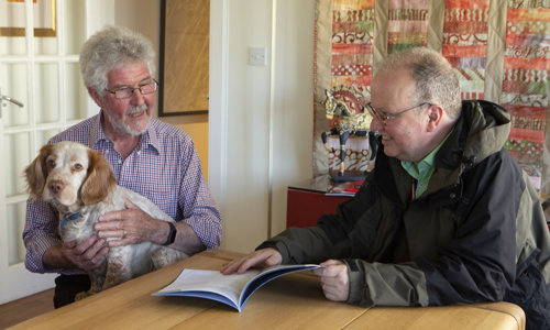 Two men at a table looking at a leaflet. One man has a dog on his lap