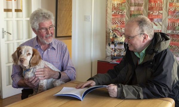 Two men at a table looking at a leaflet. One man has a dog on his lap