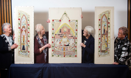 Four women stand beside carefully embroidered tapestries designed like stained glass windows