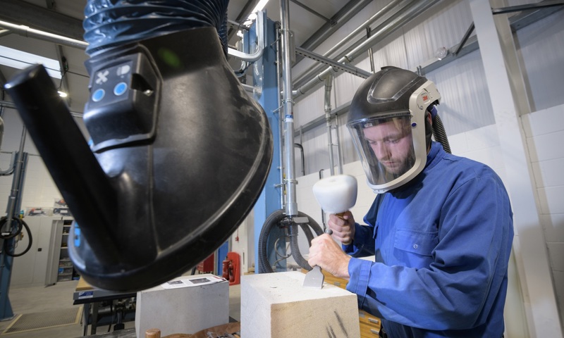 A person stonecarving wearing a full safety helmet inside a workshop