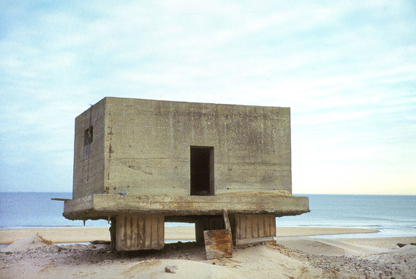 A concrete block sitting high above the sand, with the base and supporting pillars exposed