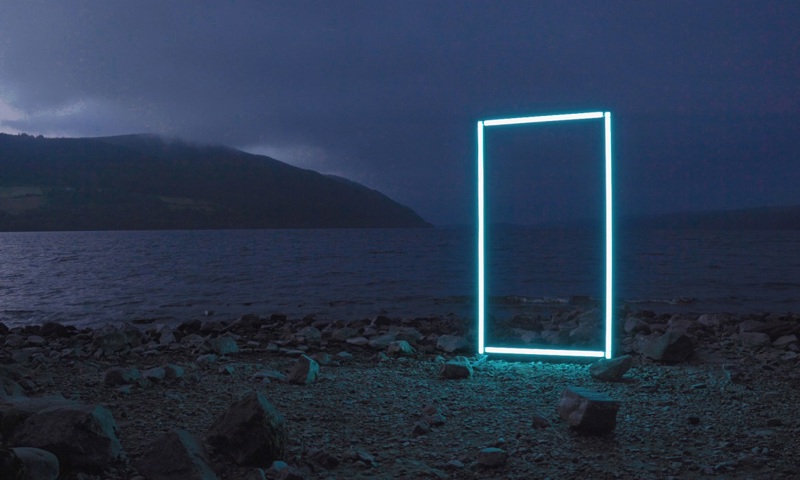 A projection of a bright blue rectangular shape, perhaps a picture frame, on the rocky shore of a loch